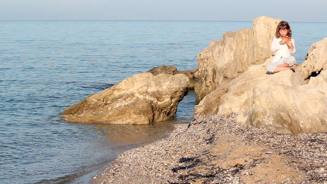 little girl sitting on a rock by the sea and playing pan pipe