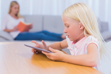 Little girl using tablet while her mother is reading on the couc