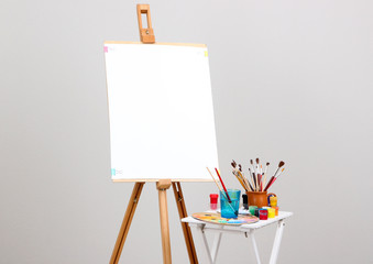 Wooden easel with clean paper and art supplies in room - obrazy, fototapety, plakaty