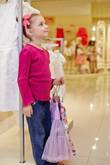 Little girl stands leaning on rack and holds hangers