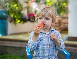 Adorable little toddler boy with blond hairs eating watermelon i