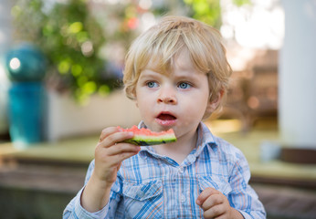 Adorable little toddler boy with blond hairs eating watermelon i