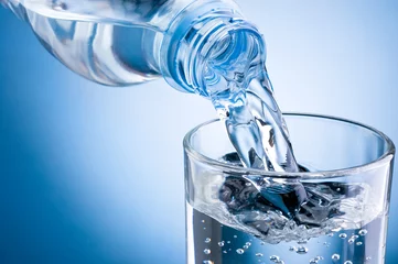 Abwaschbare Fototapete Wasser Gießen von Wasser aus der Flasche in Glas auf blauem Hintergrund