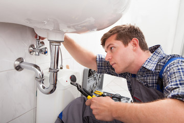 Young plumber fixing a sink in bathroom - obrazy, fototapety, plakaty