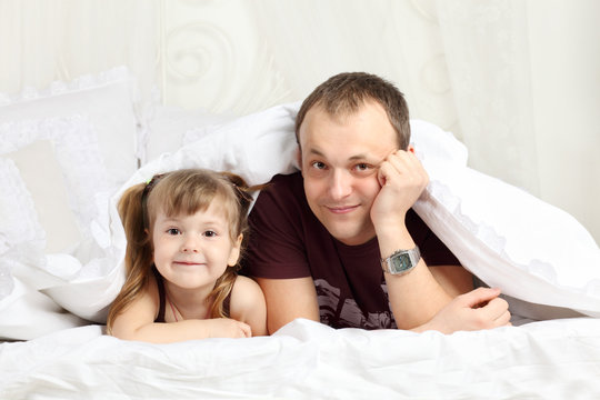 Little girl and her father look out from under blanket on bed an