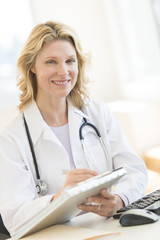 Female Doctor With Clipboard Sitting At Desk In Clinic