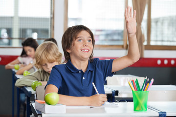 Schoolboy Raising Hand While Sitting At Desk