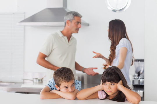 Unhappy siblings sitting in kitchen with their parents who are f