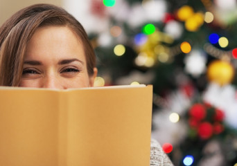 Happy young woman hiding behind book near christmas tree