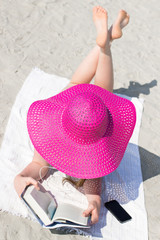 Young woman with big pink hat reading on the beach