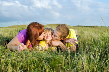 Fototapeta premium Parents kissing their daughter lying on a green meadow
