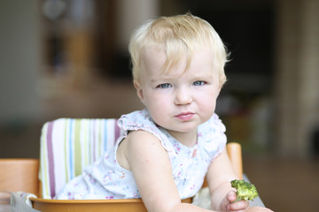 Fototapeta premium Beautiful baby girl sits in feeding chair holds steamed broccoli