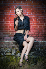 Charming young woman in black dress sitting on brick wall 