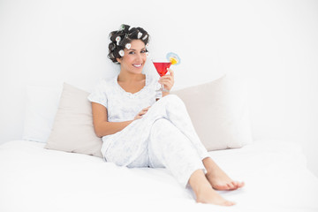 Smiling brunette in hair curlers sitting on her bed holding a co