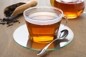 Transparent cup of black tea and teapot on wooden background