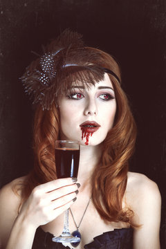 Redhead vampire woman with glass of blood. Photo in vintage styl