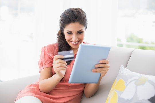 Happy brunette sitting on her sofa using tablet to shop online