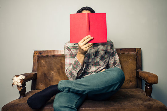 Man Hiding His Face Behind Book On Old Sofa