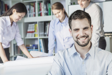 Fototapeta premium Smiling architect looking at camera while colleagues are planning around a table and looking at a blueprint