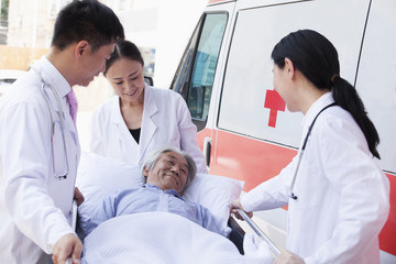 Three doctors wheeling in a elderly patient on a stretcher in front of an ambulance © xixinxing