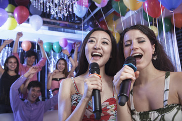 Two friends holding microphones and singing together at karaoke, friends in the background - obrazy, fototapety, plakaty