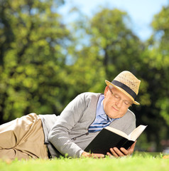 Senior man with hat lying on a grass reading a book in a park