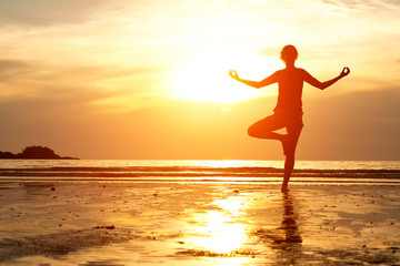 Fototapeta premium Backlit, young woman practicing yoga on the beach at sunset.