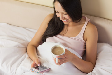 Cheerful young asian woman sitting in bed texting on her smartph