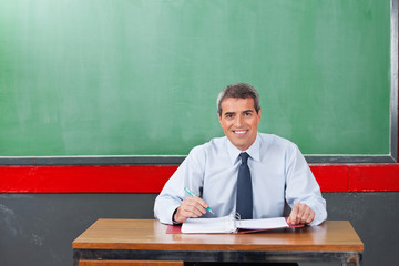 Confident Male Teacher With Pen And Binder Sitting At Desk