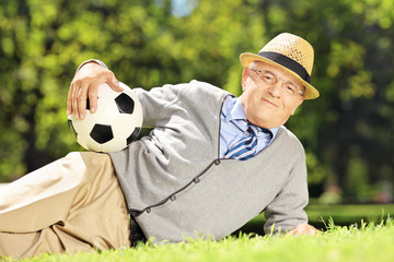 Naklejka premium Senior man with hat lying on a grass holding a ball in a park