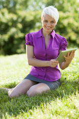 Smiling woman with tablet pc is sitting on the grass while smili