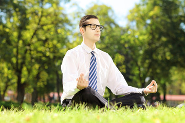 Young businessperson with eyeglasses meditating seated in park - obrazy, fototapety, plakaty