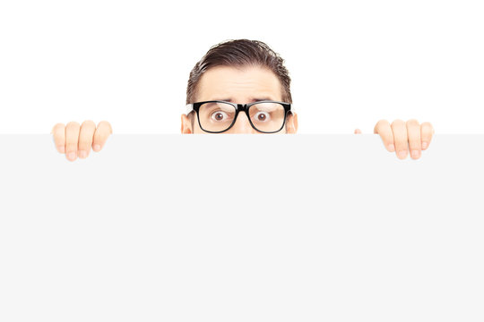 Scared young man with glasses hiding behind a blank panel