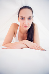 Pretty brunette lying under the sheets smiling at camera