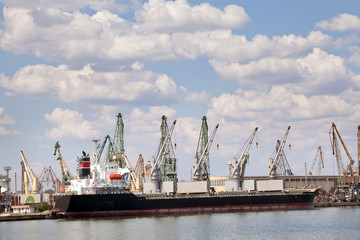 Fototapeta premium Large cargo ship in a dock at port. Cloudy sky