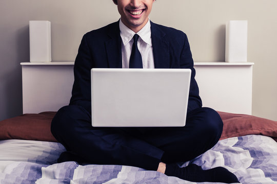 Happy young businessman sitting in bed with laptop