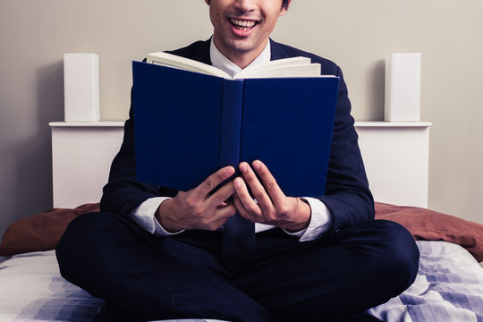 Happy young businessman sitting in bed reading book