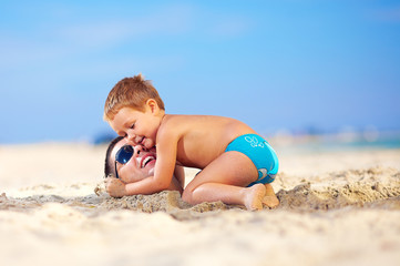 happy kid gently hugging father's head in sand on the beach