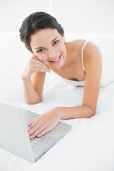 Obraz na płótnie Canvas Smiling casual brunette in white pajamas using a laptop