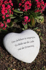 Herz mit Trauerspruch neben Blumen auf einem Grab