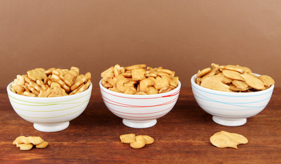 Delicious crackers in bowls on wooden table on brown background