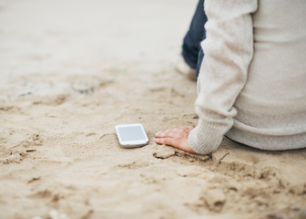 Closeup on cell phone near woman in sweater sitting on beach