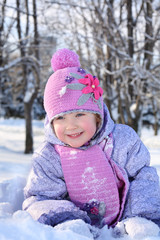 Happy little girl in pink scarf and hat lies in snow and looks