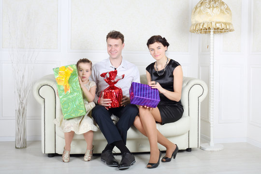 Father, mother and daughter sit at white sofa with bright gifts
