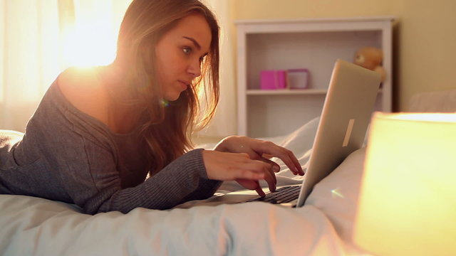 Pretty happy brunette lying on bed typing on laptop