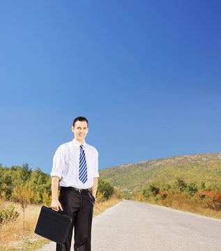 Young smiling businessperson holding a suitcase on a road