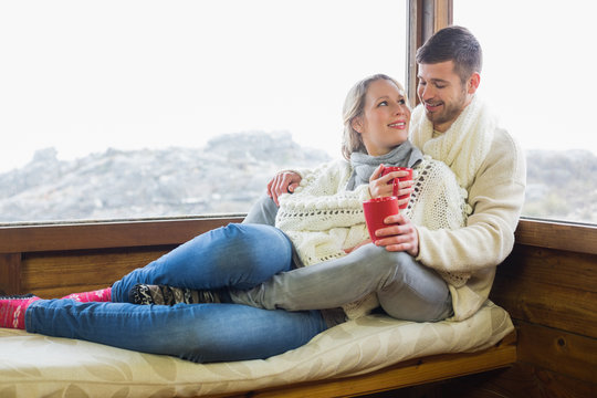 Loving couple in winter wear with cups against cabin window