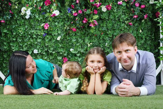 Happy family of four lie on grass near hedge with flowers