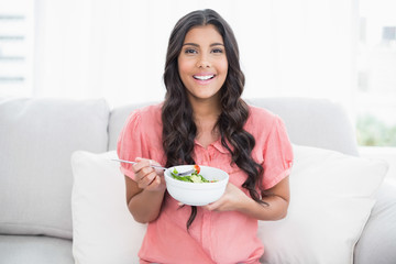 Obraz na płótnie Canvas Happy cute brunette sitting on couch holding salad bowl