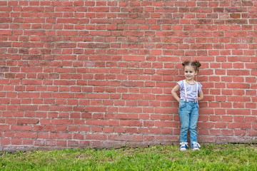 Little girl in jeans with suspenders stands near old brick wall